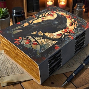 May include: A decorative book with a raven illustration on the cover, featuring a black raven perched on a branch with red and gold accents. The book has aged, gold-colored pages and a dark gray spine. A pen rests on a paper next to the book.