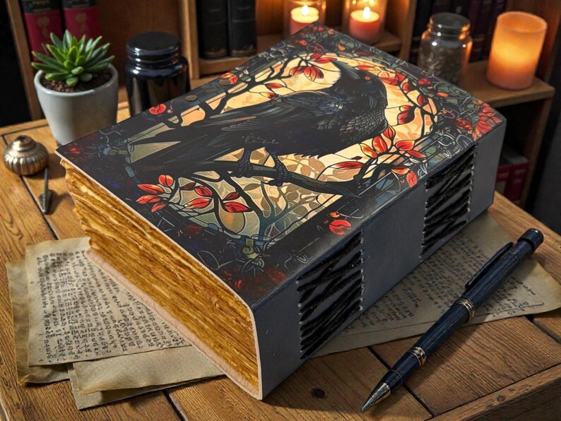 May include: A decorative book with a raven illustration on the cover, featuring a black raven perched on a branch with red and gold accents. The book has aged, gold-colored pages and a dark gray spine. A pen rests on a paper next to the book.