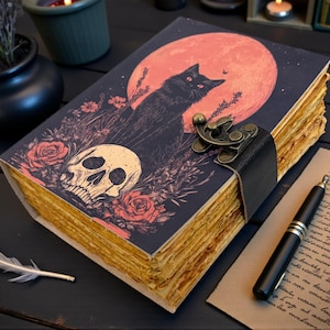 K&ouml;nnte beinhalten: Ein dekoratives Notizbuch mit einer Illustration einer schwarzen Katze vor einem roten Mond, einem Totenkopf und Rosen. Das Notizbuch hat einen Lederriemenverschluss und gealterte Papierseiten. Eine Feder und ein Stift liegen in der N&auml;he.
