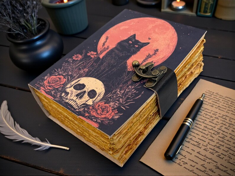 K&ouml;nnte beinhalten: Ein dekoratives Notizbuch mit einer Illustration einer schwarzen Katze vor einem roten Mond, einem Totenkopf und Rosen. Das Notizbuch hat einen Lederriemenverschluss und gealterte Papierseiten. Eine Feder und ein Stift liegen in der N&auml;he.