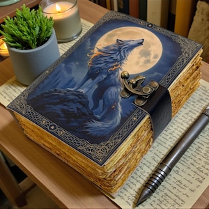 Könnte beinhalten: Ein Fantasy-Tagebuch mit einem Wolf, der den Mond auf dem Cover anheult. Das blau-goldene Cover hat ein verziertes Design und einen Verschluss. Ein Stift mit weißer Feder liegt auf einer Seite neben dem Tagebuch.