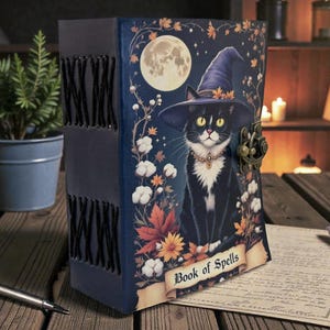 May include: A dark blue, leather-bound journal with a whimsical illustration of a black cat wearing a witch's hat, a full moon, and the words "Book of Spells." The journal has a metal clasp and decorative stitching along the spine.