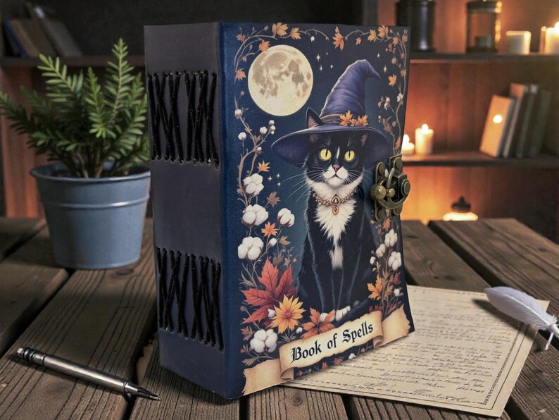 May include: A dark blue, leather-bound journal with a whimsical illustration of a black cat wearing a witch's hat, a full moon, and the words "Book of Spells." The journal has a metal clasp and decorative stitching along the spine.