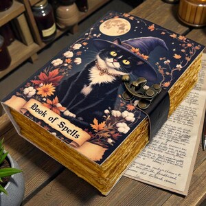 May include: A decorative book titled "Book of Spells" featuring a black and white cat wearing a witch hat. The book has a dark blue cover with floral and moon illustrations, a leather clasp, and aged-looking pages.