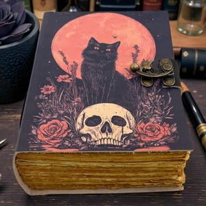 K&ouml;nnte beinhalten: Dekoratives Notizbuch mit einer Illustration einer schwarzen Katze, einem Totenkopf und Rosen vor einem roten Mond. Das Notizbuch hat einen Metallverschluss und gealterte Seiten. Das Cover ist dunkel mit Vintage-&Auml;sthetik.