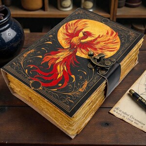 Peut inclure: Un journal de style vintage avec une couverture noire orn&eacute;e d'un ph&eacute;nix rouge et jaune vif sur fond de lune jaune. Le journal est dot&eacute; d'une fermeture &agrave; lani&egrave;re en cuir et de pages en papier vieilli.
