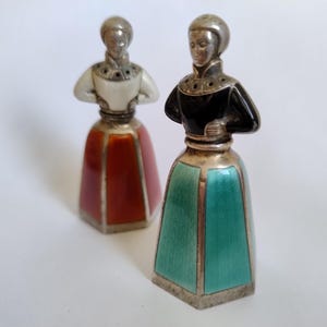 May include: Two vintage salt and pepper shakers. Each shaker is shaped like a woman in a dress. One has a red skirt and white top, the other has a teal skirt and black top. Both have silver-colored heads and accents.