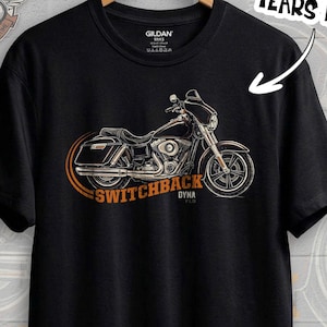 Puede incluir: Camiseta negra con un diseño gráfico de motocicleta y las palabras "SWITCHBACK DYNA" en naranja y blanco. La camiseta también tiene el texto "ALL FACTORY COLORS YEARS INSIDE" y "FAST DELIVERY". La camiseta está colgada en una percha de madera.