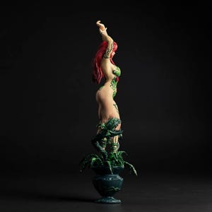 May include: A figurine of a woman with red hair and green vine-like body art. The figure is standing on a dark green planter with a plant. The background is a solid black.