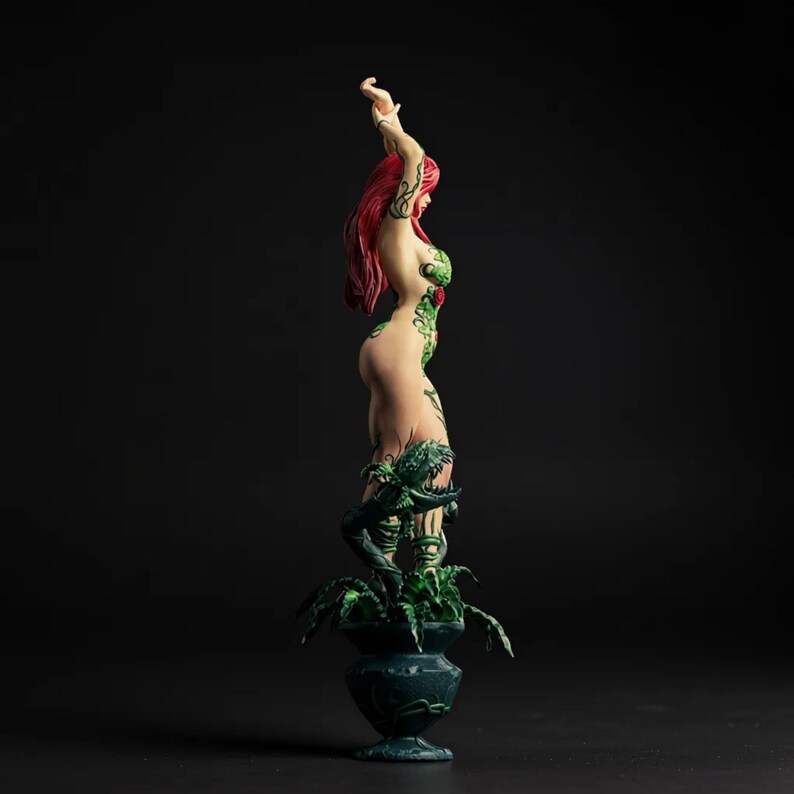 May include: A figurine of a woman with red hair and green vine-like body art. The figure is standing on a dark green planter with a plant. The background is a solid black.
