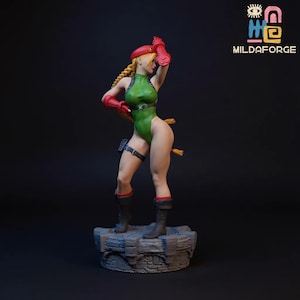 Puede incluir: Figura detallada de un personaje femenino con un body verde, guantes rojos y boina roja. La figura tiene el pelo rubio en una trenza y posa sobre una base de piedra. La figura lleva botas negras con ribetes rojos. El logotipo "MILDAFORGE" está en la esquina superior derecha.