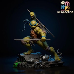 May include: A detailed figurine of Leonardo from Teenage Mutant Ninja Turtles in a dynamic pose, wielding a katana. The figure is predominantly green and brown, with blue accents. The base is a textured, grey surface with green and silver details. The logo "MILDAFORGE" is visible.
