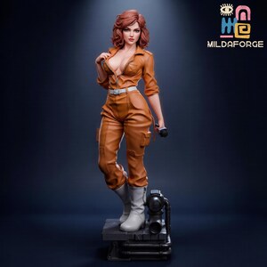 Può includere: Statuetta dettagliata di una donna in tuta arancione, stivali bianchi e cintura bianca. La figura ha i capelli rossi e tiene in mano un microfono nero. La base è una piattaforma grigio scuro con tubi. Il logo "MILDAFORGE" è in alto a destra.