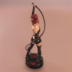 May include: A figurine of a woman with red hair, dressed in a black outfit, holding a whip. The figure stands on a round, black base with red accents. The background is a gradient of pink and peach.