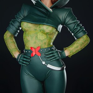 Pode incluir: Um traje de super-her&oacute;i verde com uma jaqueta curta, luvas e cal&ccedil;as. O corpete tem um padr&atilde;o floral verde claro, e o cinto tem uma fivela vermelha "X". O traje est&aacute; em um fundo preto.