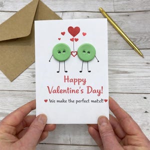May include: A Valentine's Day card with a white background, featuring two green buttons with drawn arms and legs, connected by a heart. The card reads "Happy Valentine's Day!" and "We make the perfect match!" in red.
