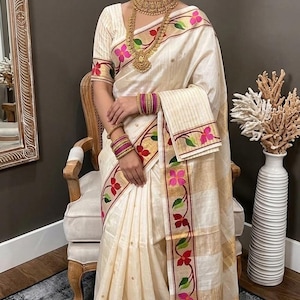 Peut inclure: Un sari blanc cassé avec des rayures horizontales dorées et une bordure décorative ornée de motifs floraux roses et rouges avec des feuilles vertes. La tenue est accompagnée de bijoux dorés, dont un collier, des boucles d'oreilles et des bracelets.