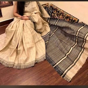 Può includere: Un sari beige caratterizzato da un tessuto testurizzato con strisce orizzontali marrone scuro e nere lungo il bordo e il pallu. L'indumento presenta un aspetto naturale e intrecciato con una tavolozza di colori coerente in tutte le sezioni.