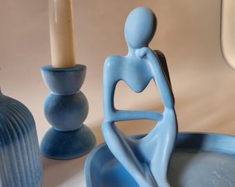 May include: A light blue abstract figurine of a seated person resting their chin on one hand, positioned on a matching light blue tray. Next to the figurine is a light blue candle holder with a white taper candle and a ribbed blue vase.