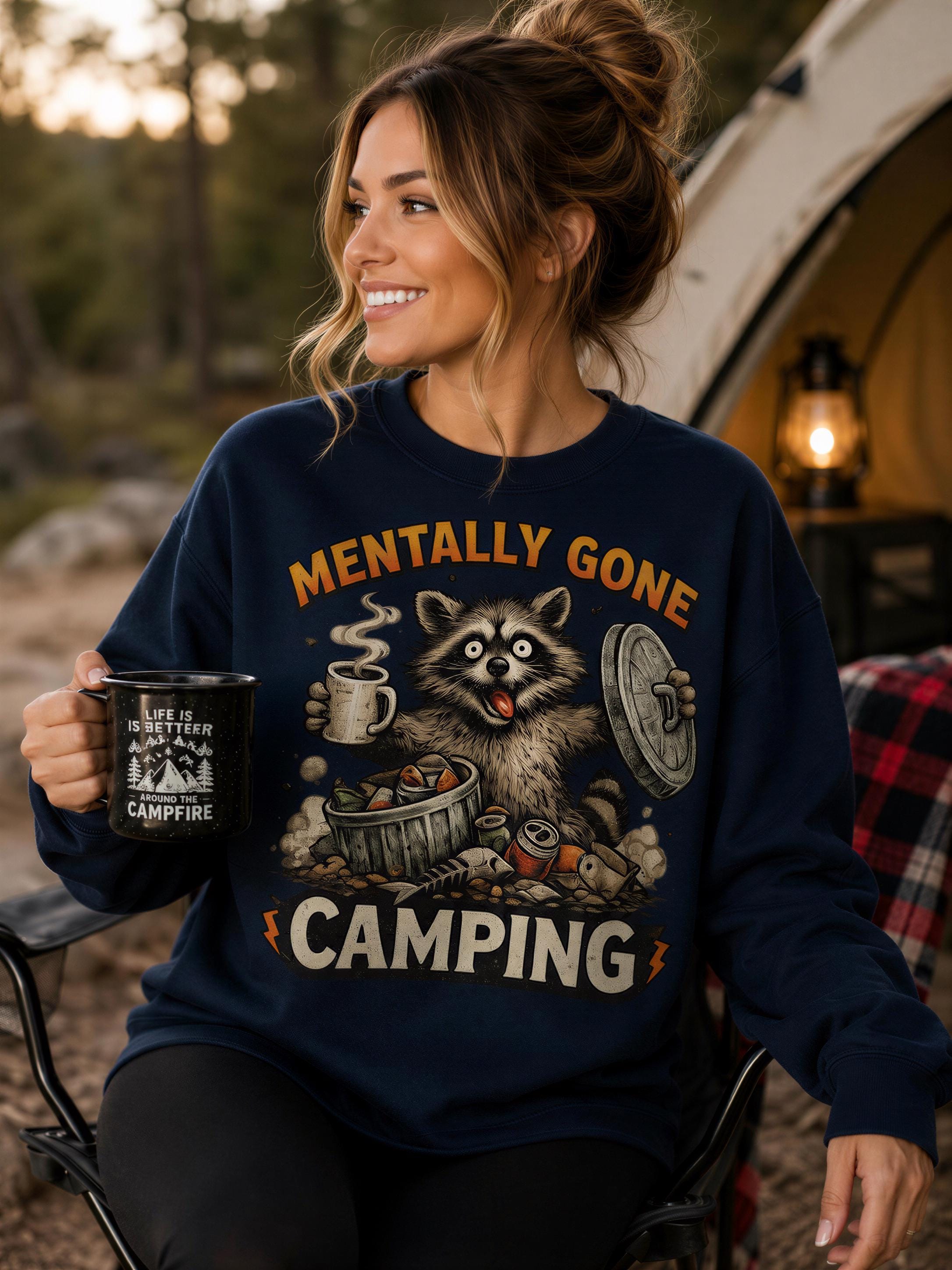 A woman wearing a blue sweatshirt with a raccoon graphic and the text "Mentally Gone Camping" is sitting outdoors, holding a coffee mug.