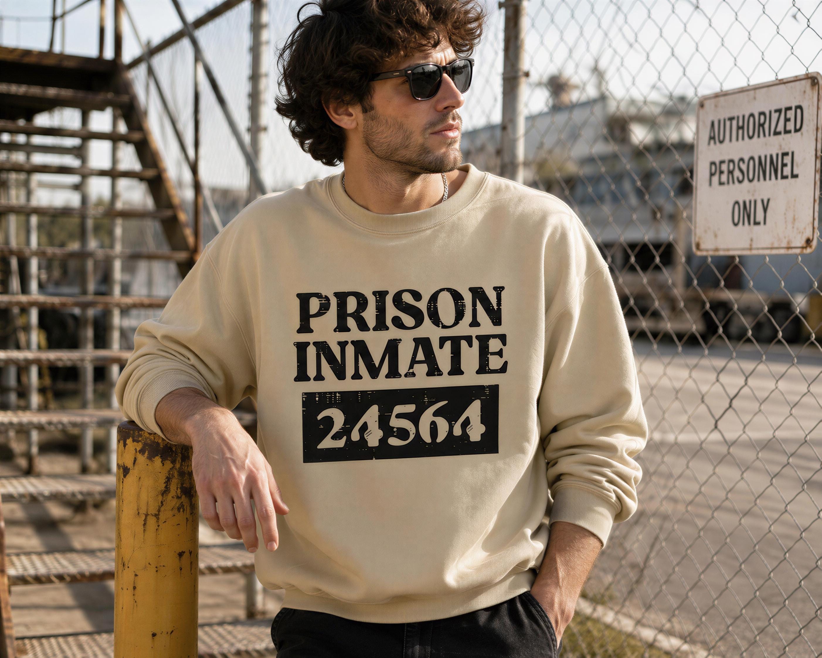 A man wearing a sweatshirt with the text "PRISON INMATE" and the number "24564" printed on it, standing in front of a chain-link fence.
