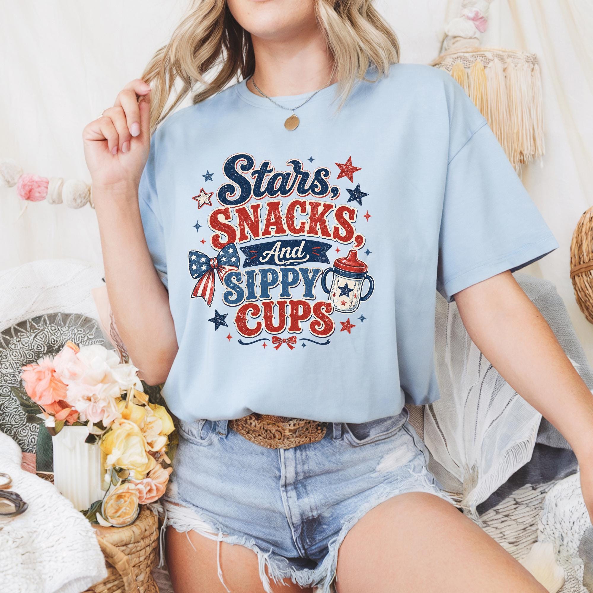 A woman wearing a light blue t-shirt with a patriotic design and denim shorts is sitting in a room with a floral arrangement.