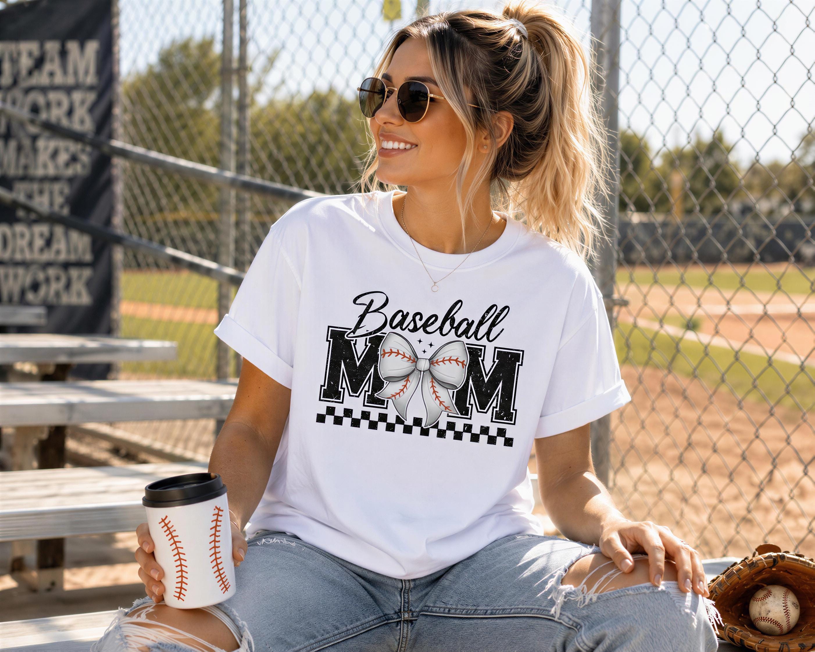 A woman wearing a white t-shirt with a baseball graphic and holding a coffee cup is sitting on a bench in a baseball field.
