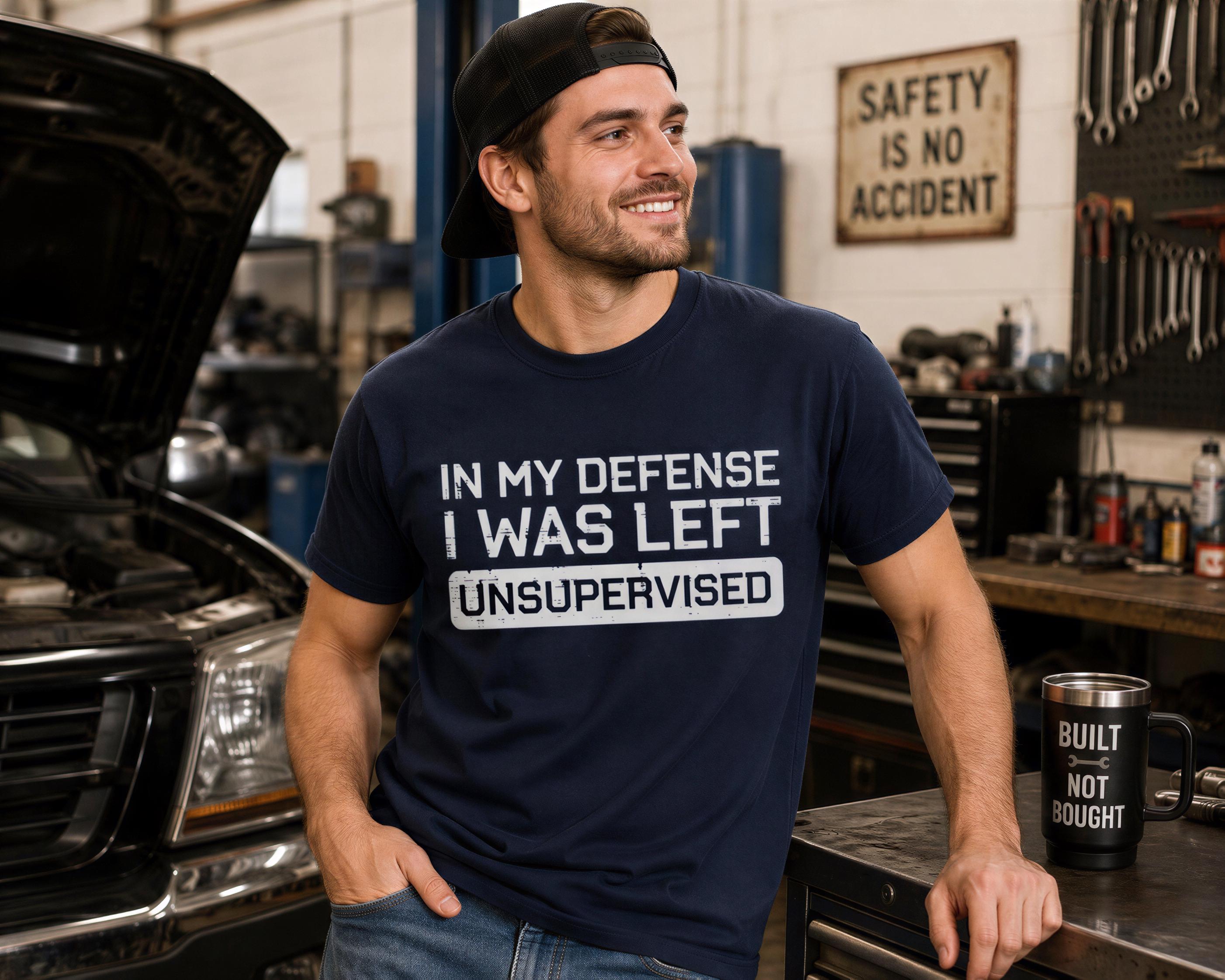 A man wearing a navy blue t-shirt with the text "IN MY DEFENSE I WAS LEFT UNSUPERVISED" is standing in a garage, smiling and leaning against a workbench.