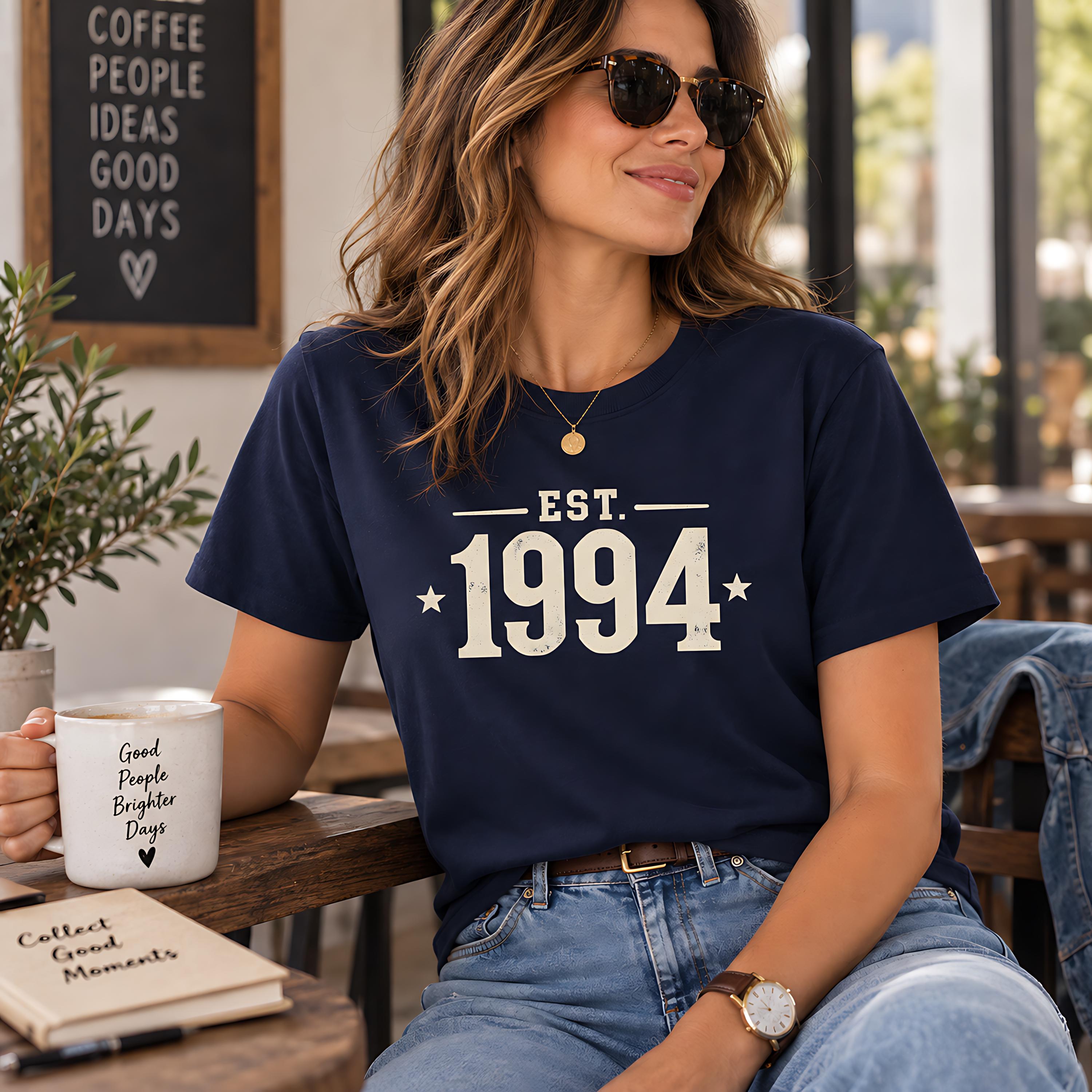 A woman wearing a navy blue t-shirt with the text "EST. 1994" and sunglasses is sitting at a table in a cafe, holding a coffee mug and reading a book.