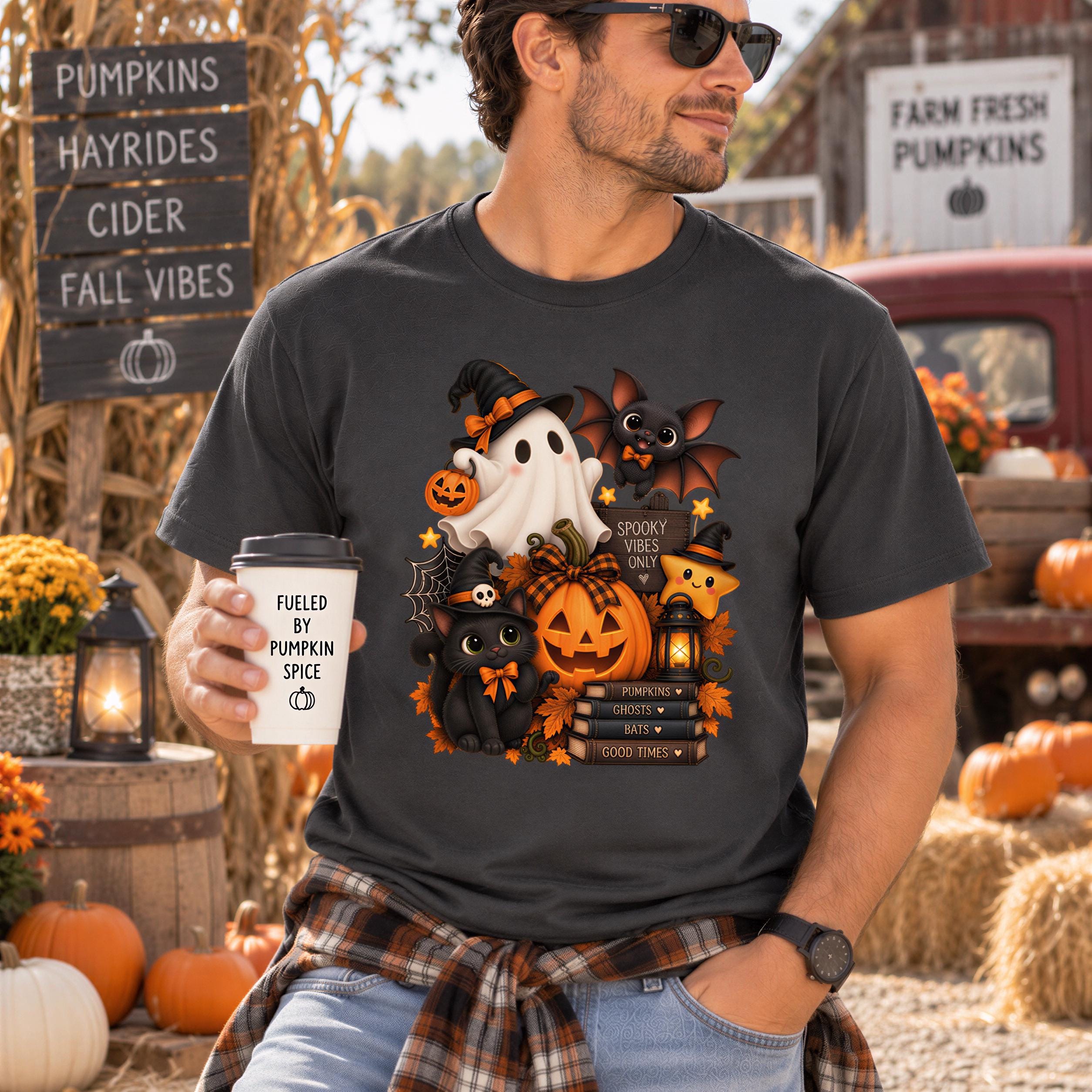 A man wearing a black t-shirt with a Halloween-themed graphic and holding a coffee cup stands in front of a sign for pumpkins, hayrides, cider, fall vibes, and a farm fresh pumpkin sign.