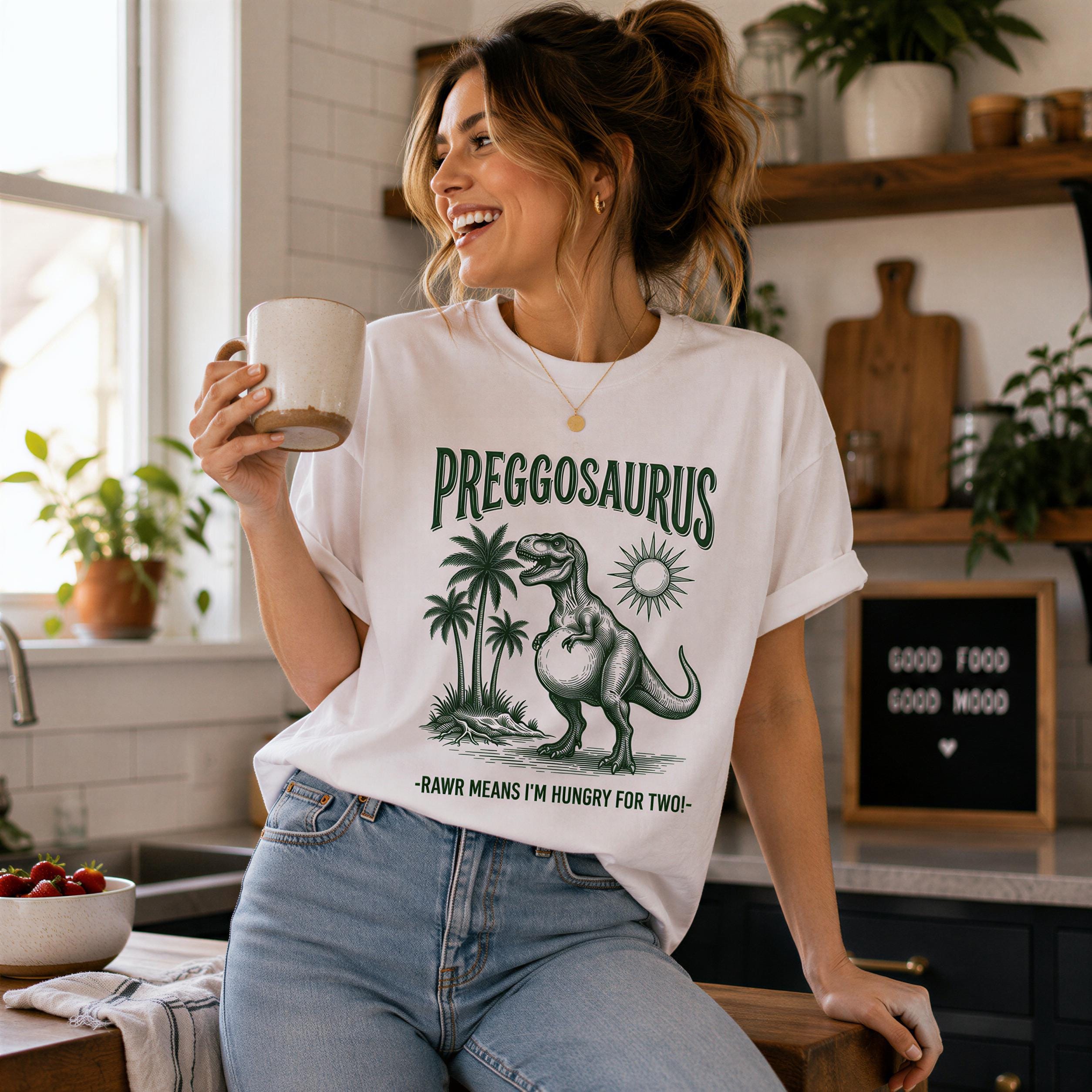 A woman wearing a white t-shirt with a graphic of a dinosaur and the text "PREGOSAURUS" is smiling and holding a cup in a kitchen setting.