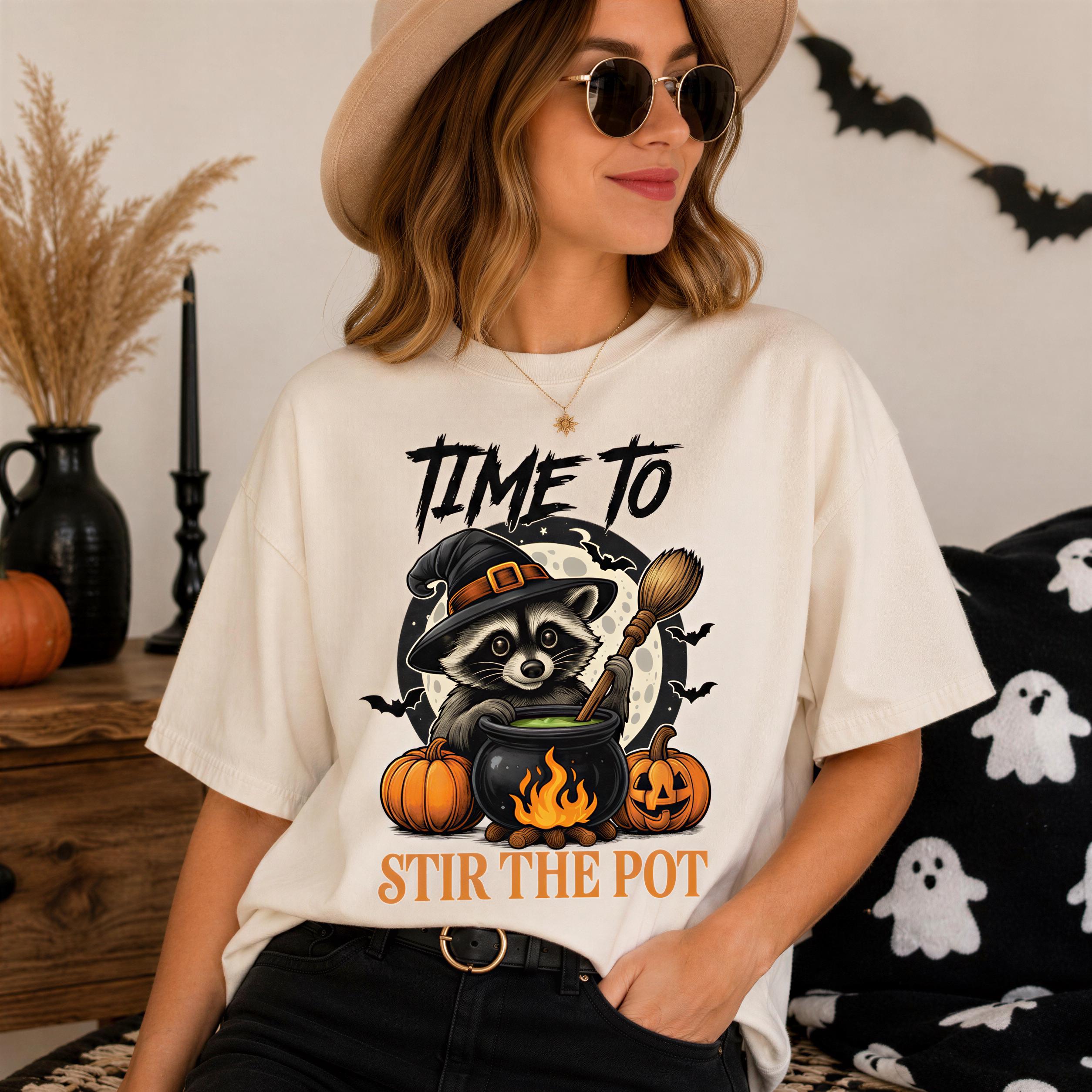 A woman wearing a white t-shirt with a graphic design of a raccoon stirring a pot, surrounded by Halloween-themed elements such as pumpkins and bats.
