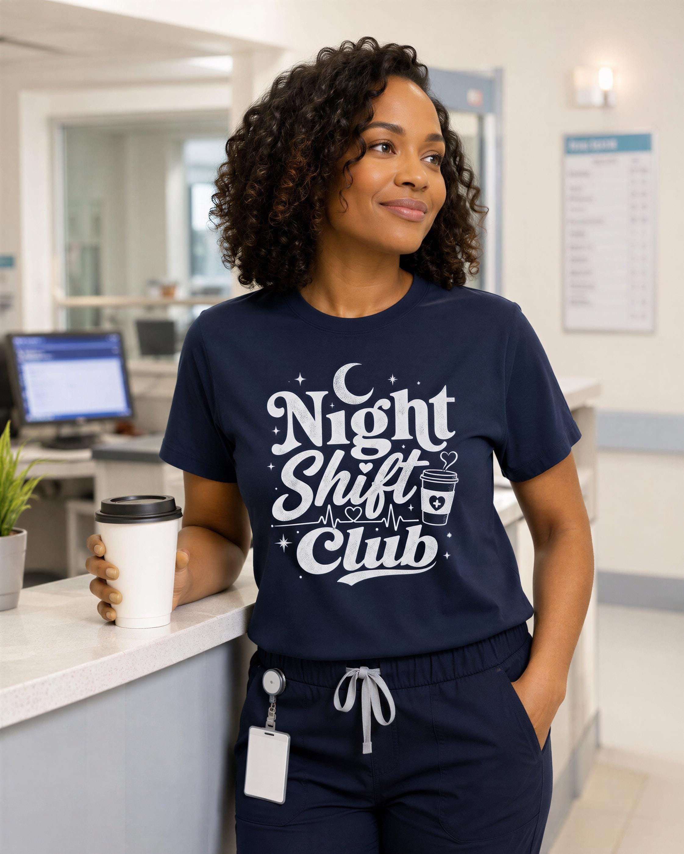 A woman wearing a navy blue t-shirt with the text "Night Shift Club" is standing in a room with a computer monitor, keyboard, and potted plant visible in the background.