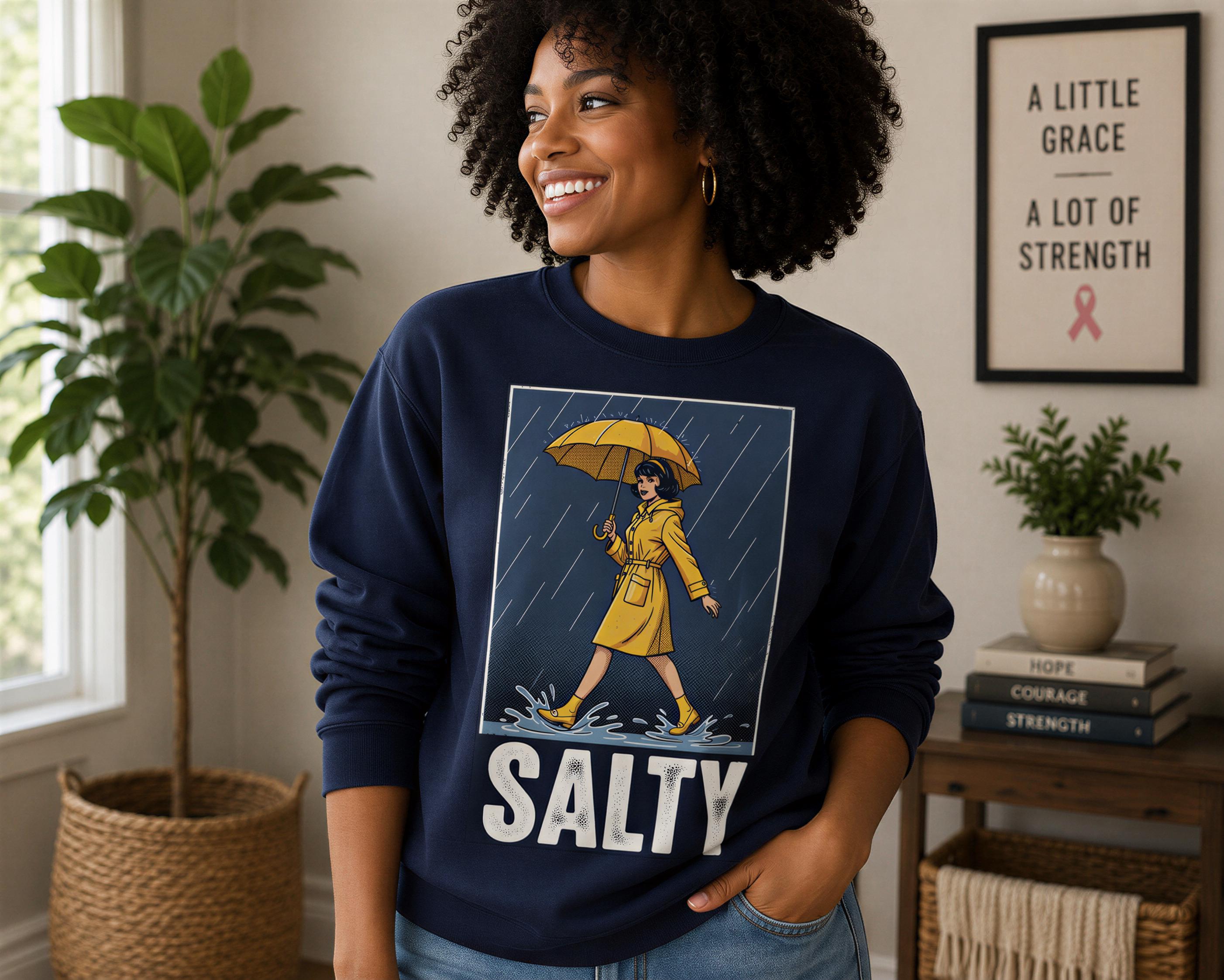 A woman wearing a navy blue sweatshirt with a graphic of a woman holding an umbrella and the word "SALTY" printed on it, smiling and looking to the side.