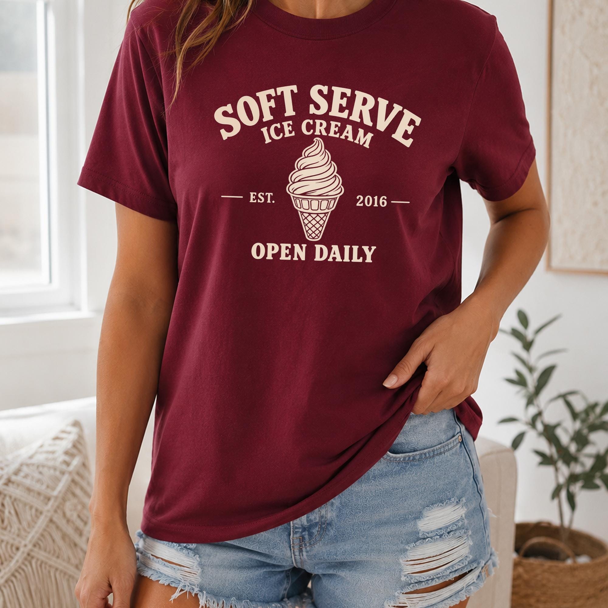 A woman wearing a burgundy t-shirt with white text and an ice cream cone graphic, standing in a room with a plant in the background.