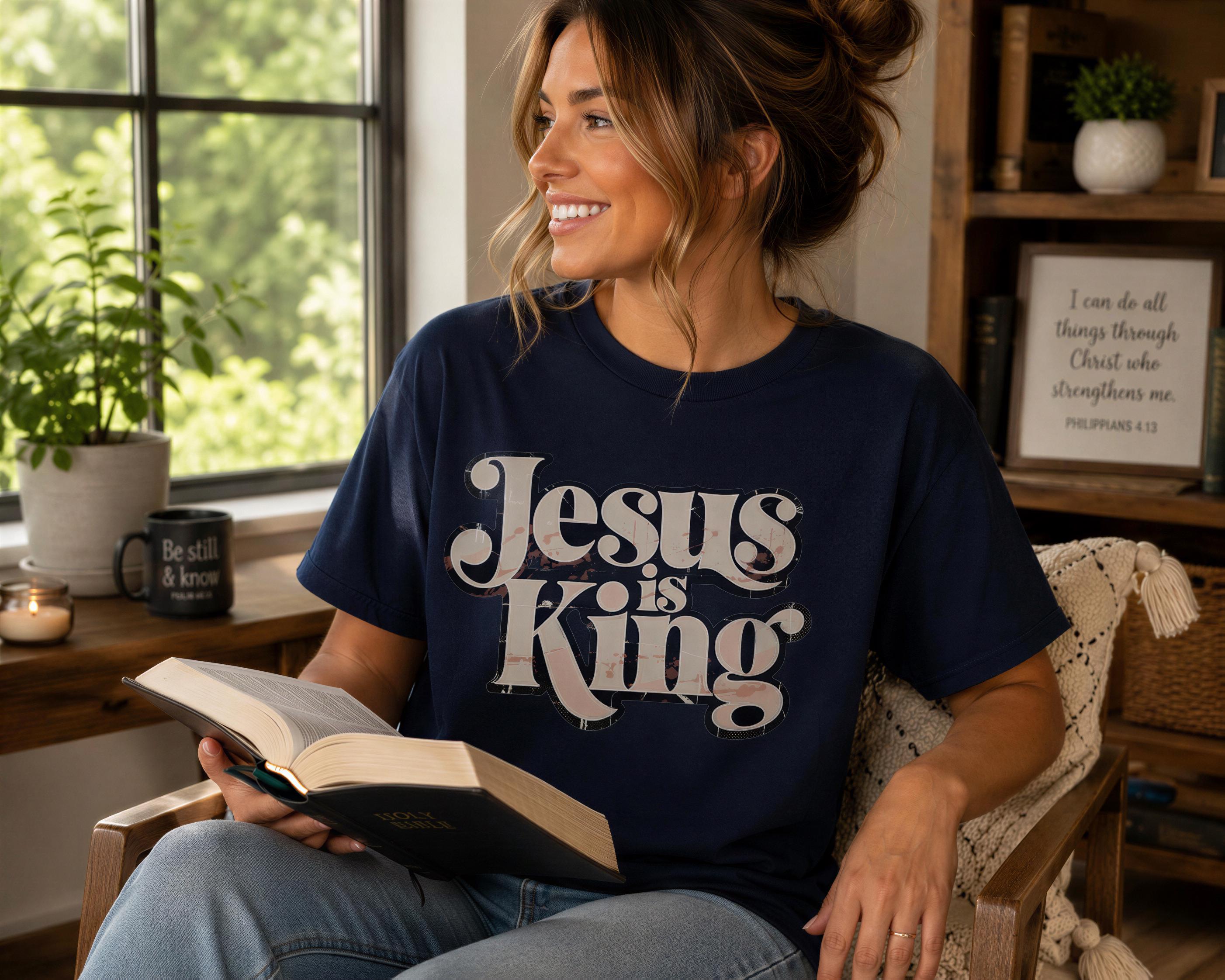 A woman wearing a blue t-shirt with the text "Jesus is King" is sitting in a chair and reading a book.