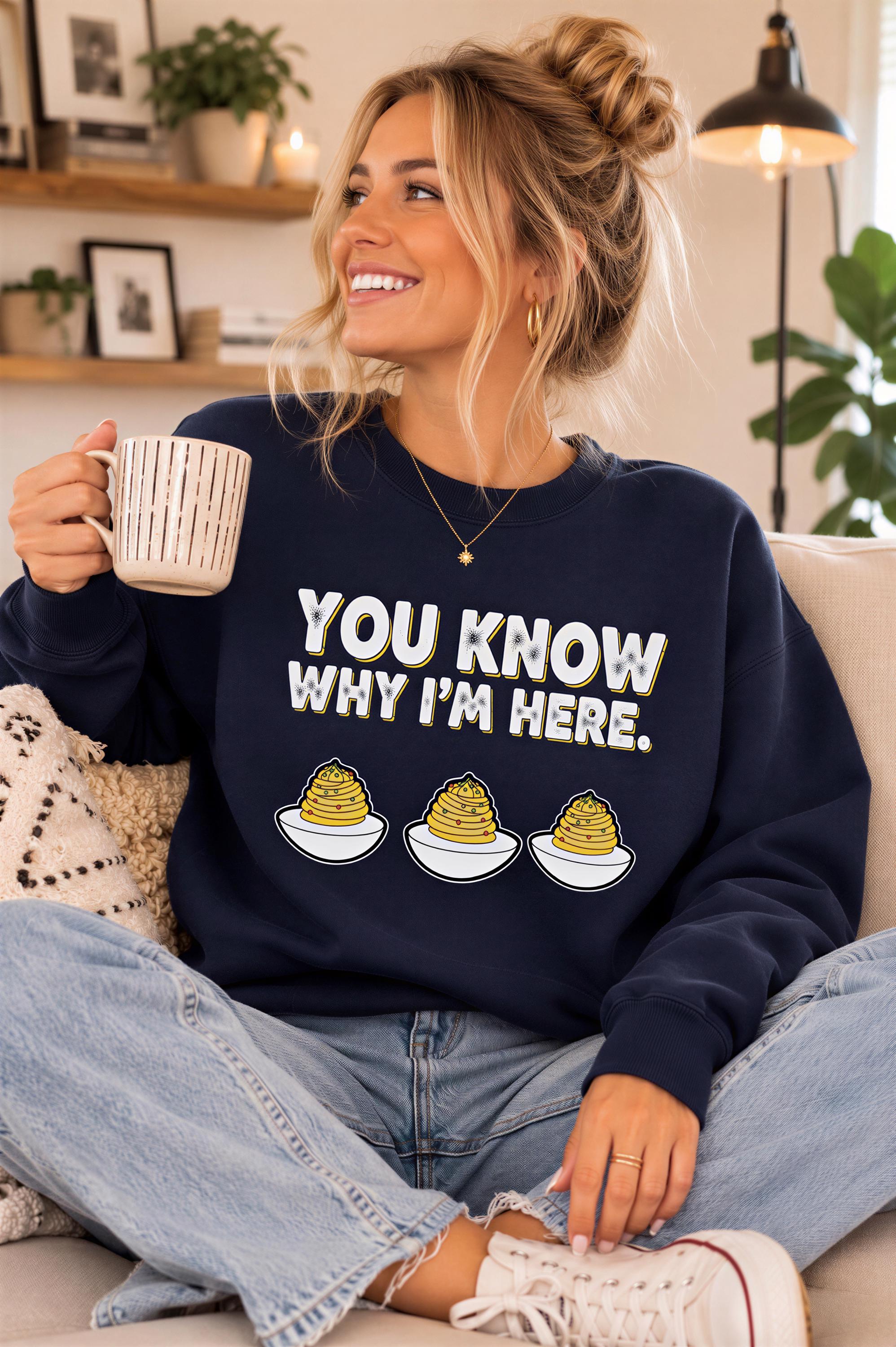 A woman sitting on a couch, smiling and holding a coffee mug, wearing a navy blue sweatshirt with a humorous text graphic.