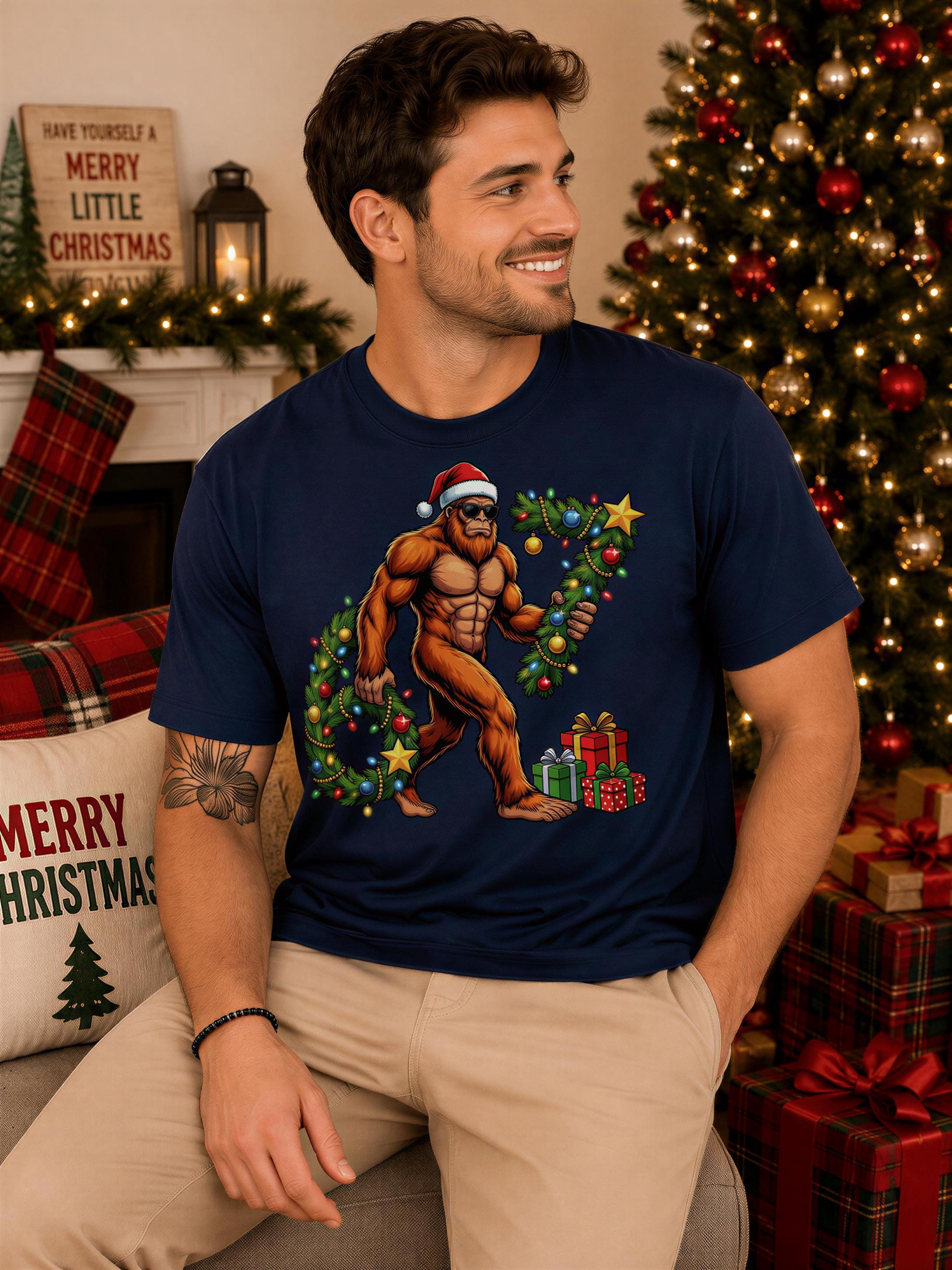 A man wearing a blue t-shirt with a cartoon image of a man in a Santa hat and holding Christmas presents is sitting in front of a Christmas tree.
