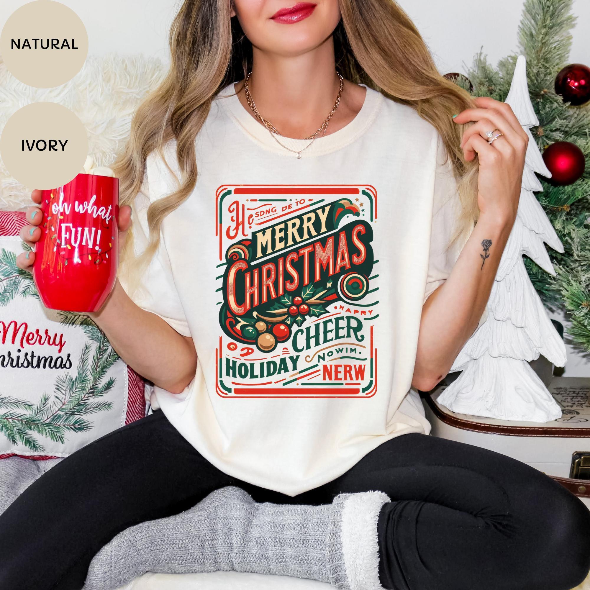 A woman wearing a white t-shirt with a Christmas-themed graphic and holding a red mug, sitting in front of a Christmas tree.