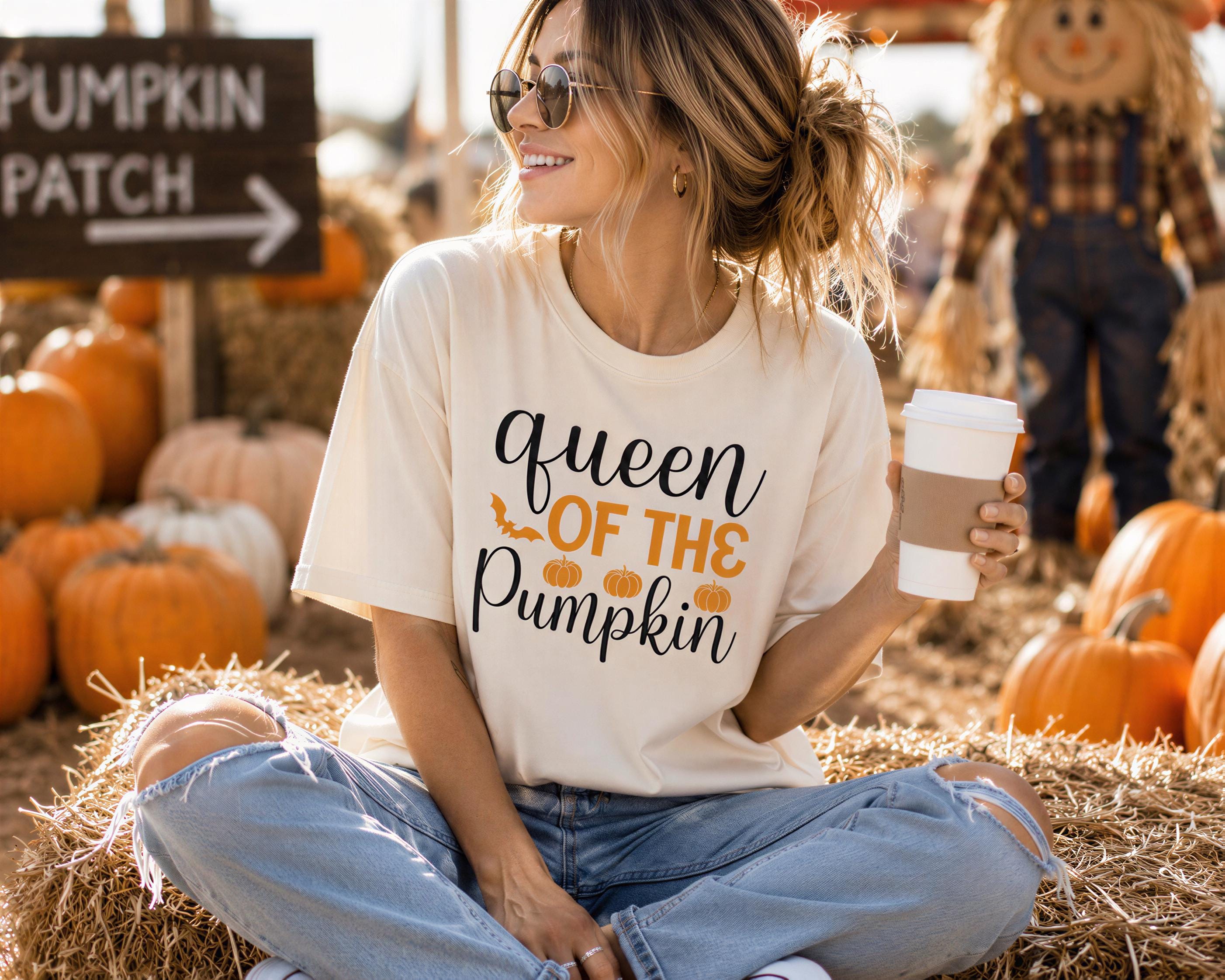 A woman sitting on hay bales in a pumpkin patch, wearing a white t-shirt with the text "queen of the pumpkin" and holding a coffee cup.