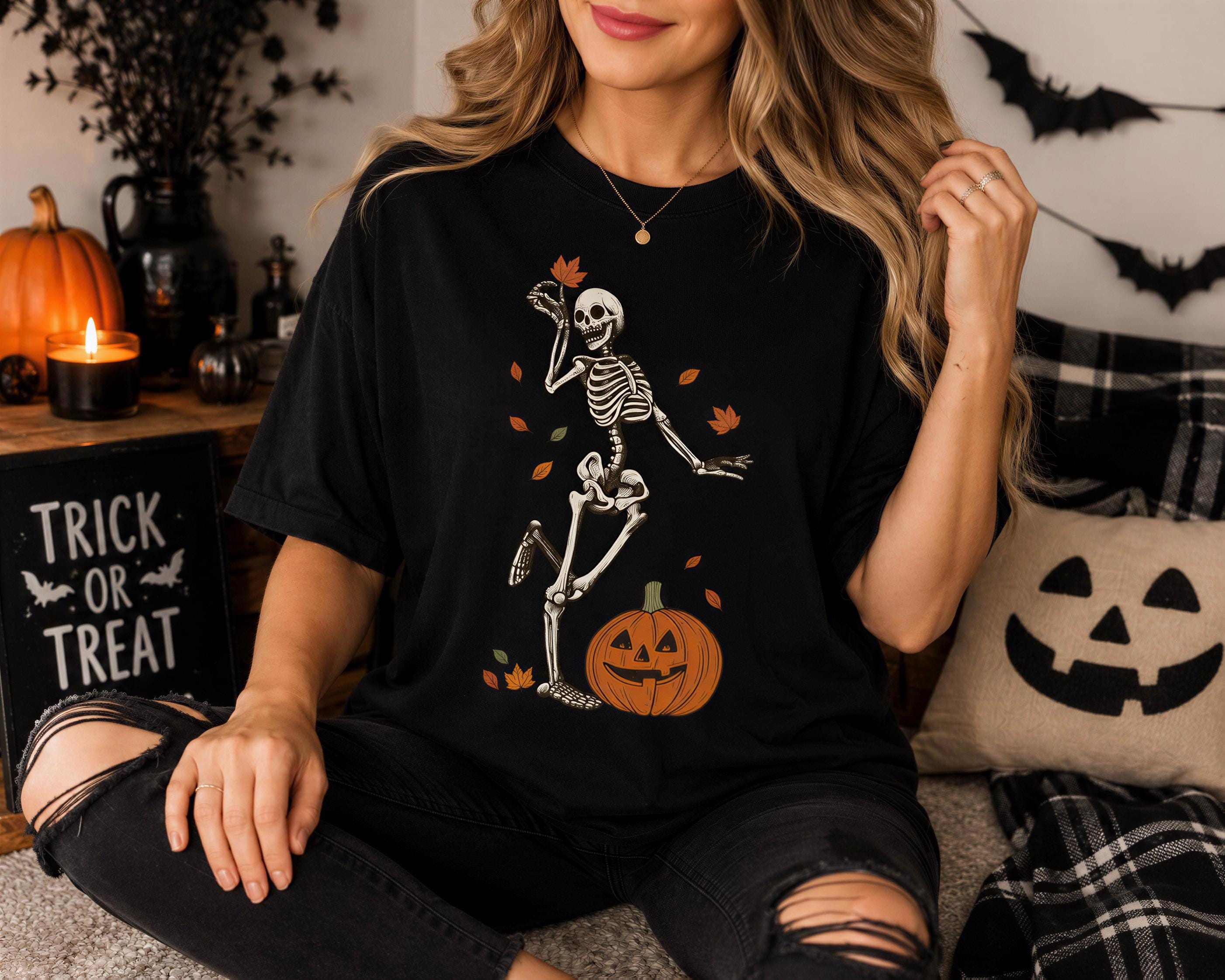 A woman wearing a black t-shirt with a skeleton and jack-o-lantern graphic is sitting on a couch with Halloween decorations in the background.