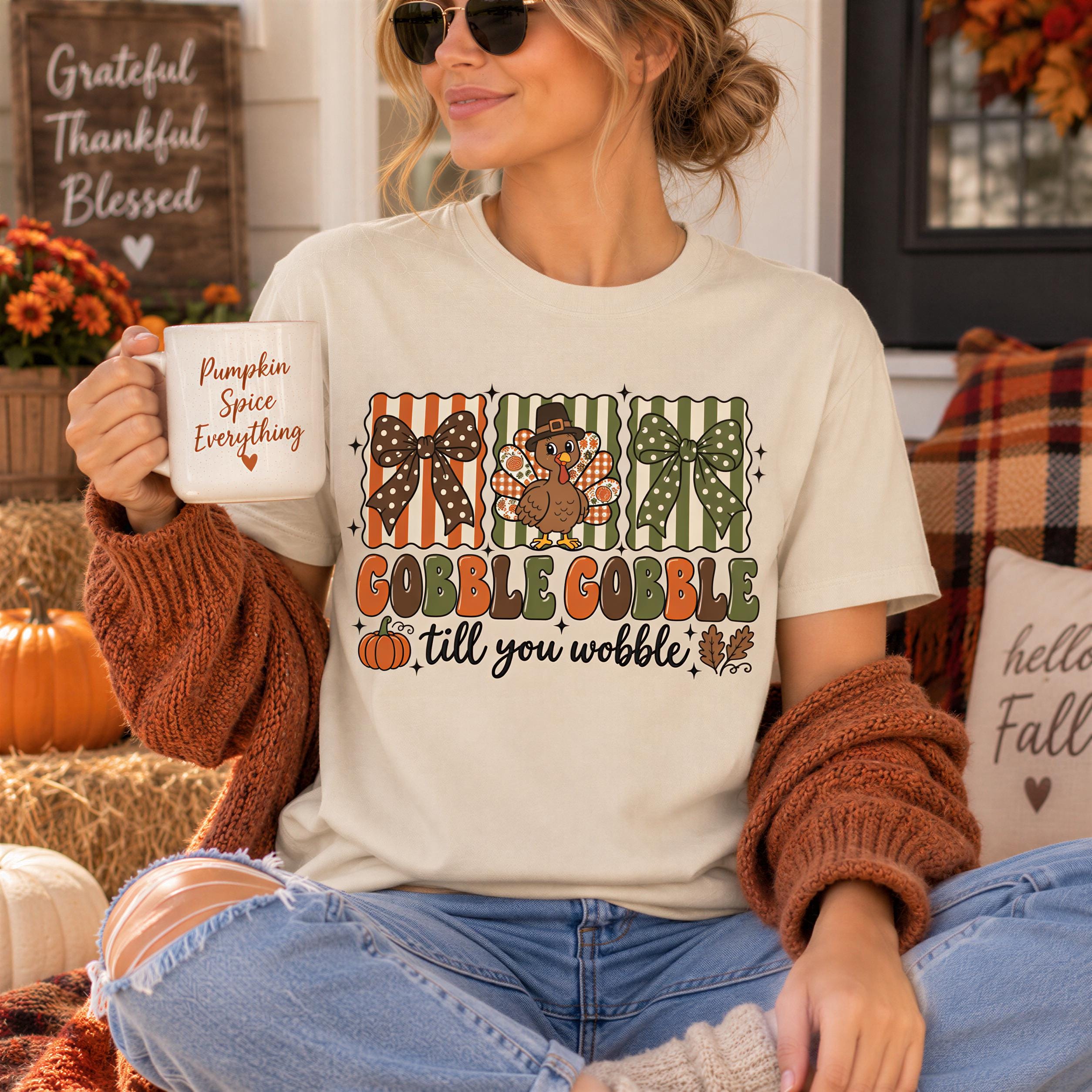 A woman wearing a white t-shirt with a graphic design of a turkey and autumn leaves, holding a coffee mug and sitting on a couch with a pumpkin and other fall decorations in the background.