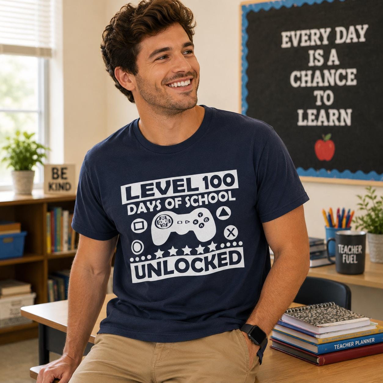 A man wearing a blue t-shirt with a graphic design and text, smiling and sitting in a classroom setting with a bulletin board and bookshelves in the background.