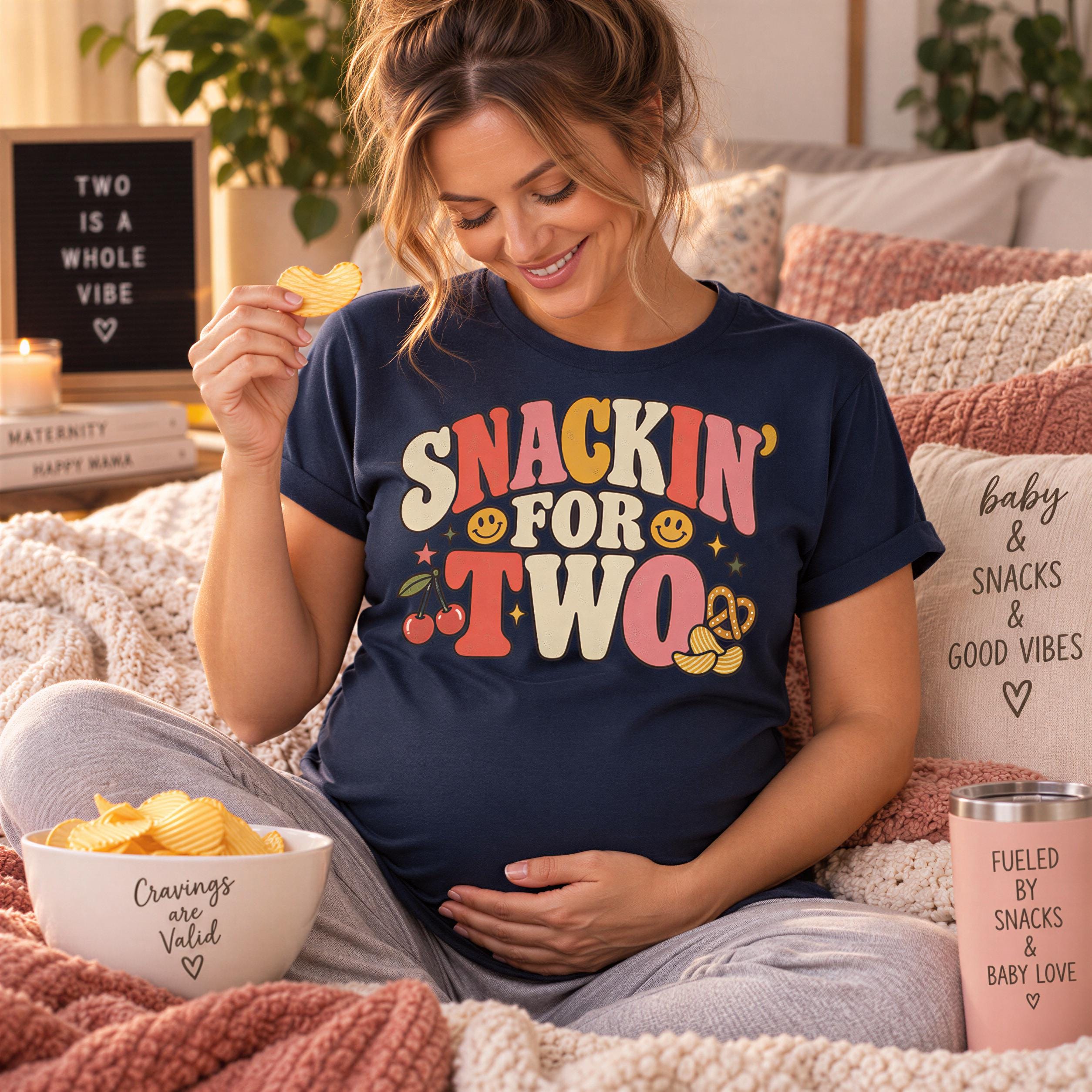 A pregnant woman is sitting on a bed, holding a snack and smiling, with a bowl of chips and a sign that says "Two is a whole vibe" in the background.