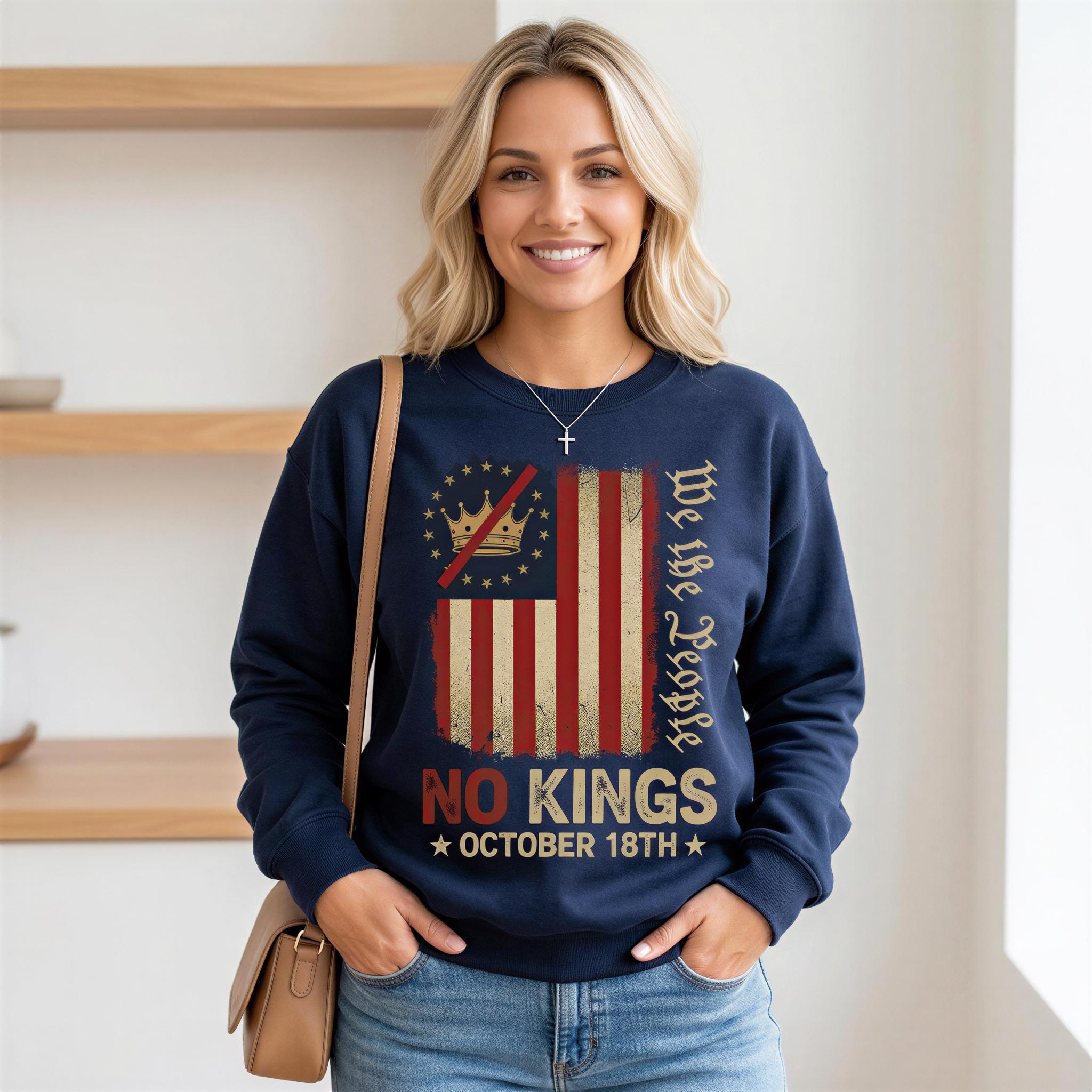A woman wearing a navy blue sweatshirt with a graphic design that says "No Kings" and "We the People" printed on it. She is smiling and standing in front of a white wall.