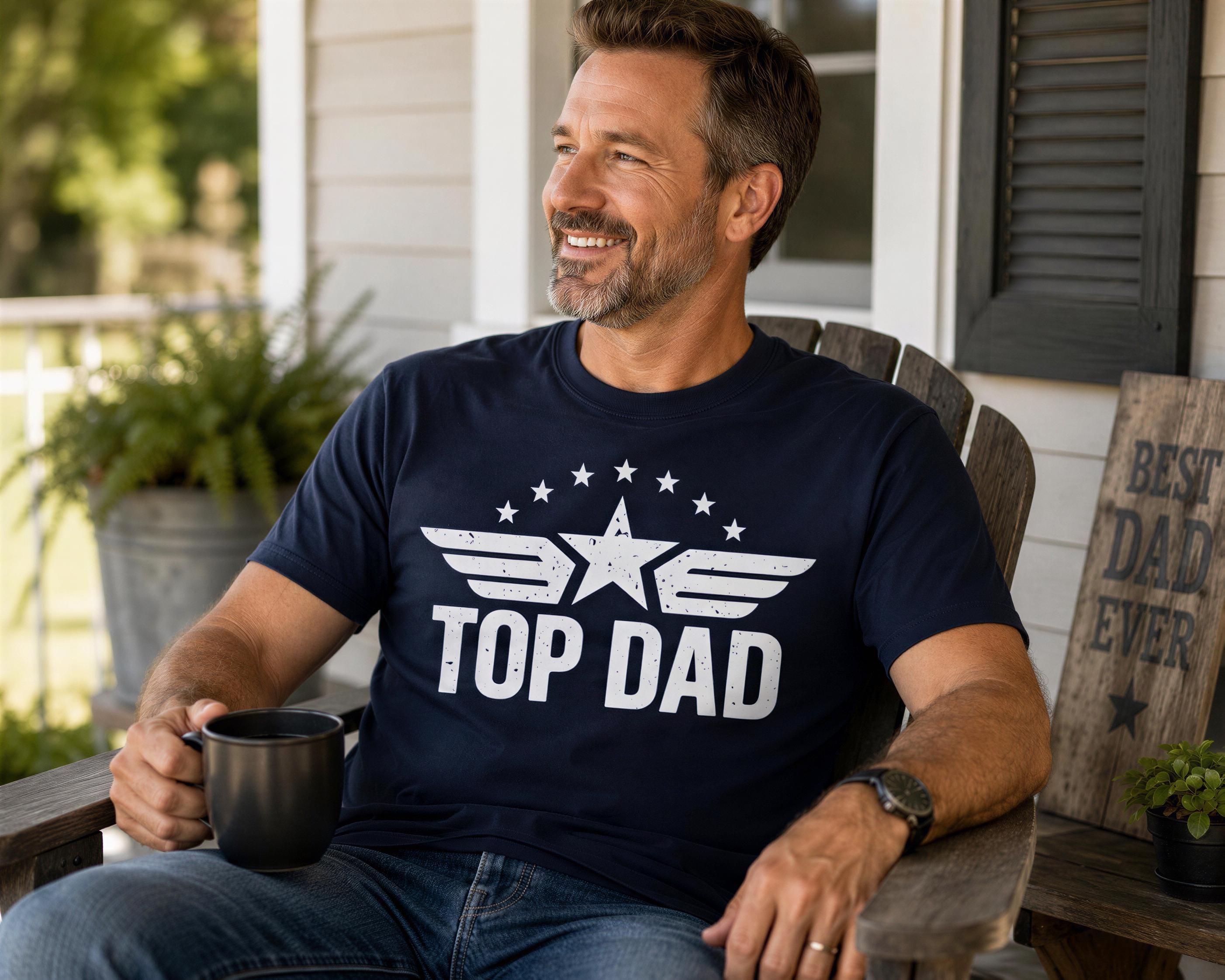 A man wearing a navy blue t-shirt with the text "TOP DAD" is sitting on a wooden bench, holding a black mug and smiling.
