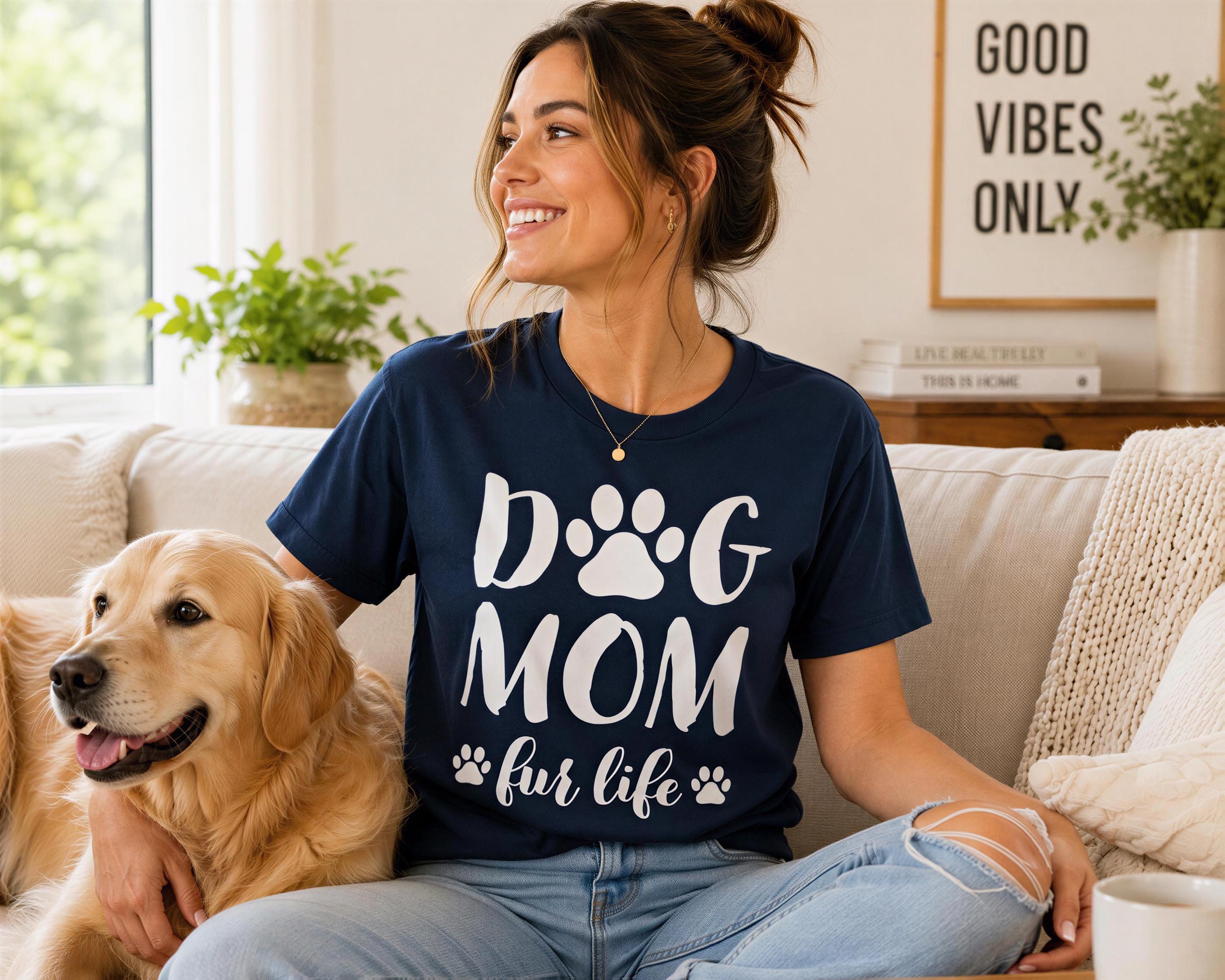 A woman wearing a navy blue t-shirt with the text "DOG MOM fur life" sits on a couch with a golden retriever dog.