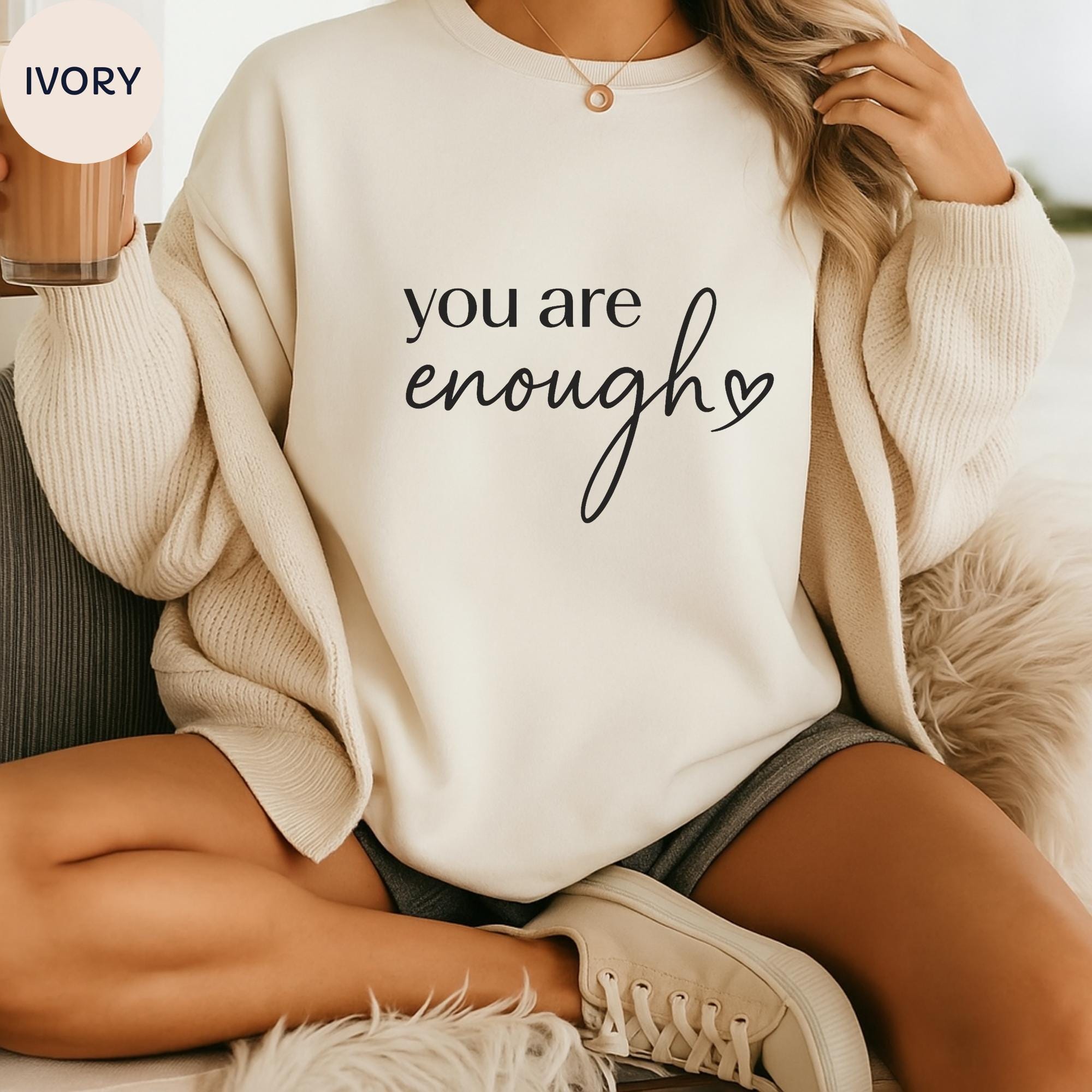 A person wearing a white sweater with the text "you are enough" printed on it, sitting on a couch with their legs crossed.