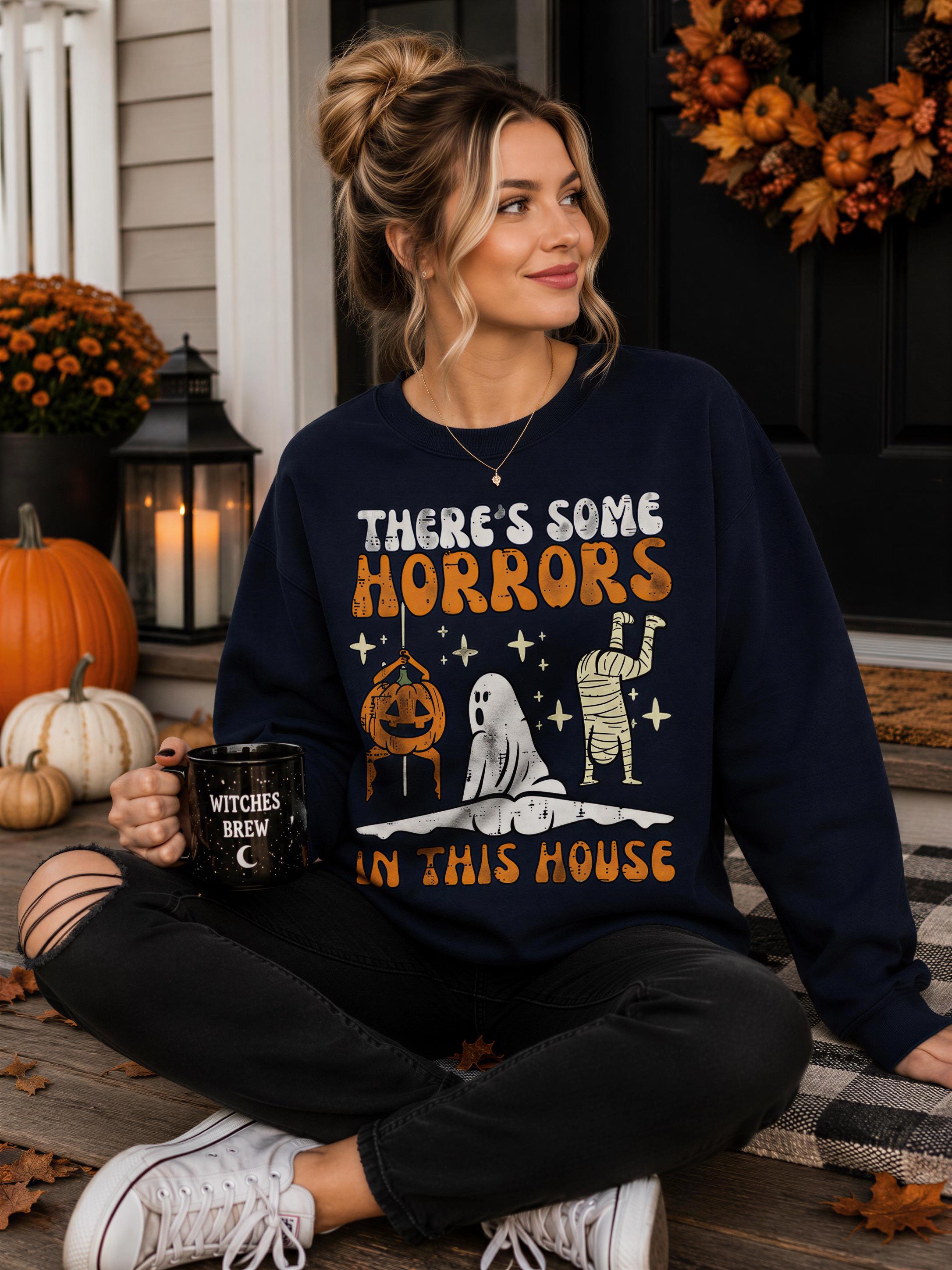A woman sitting on a porch, wearing a black sweatshirt with a Halloween-themed design, holding a mug with the text "There's some Horrors in this House" printed on it.