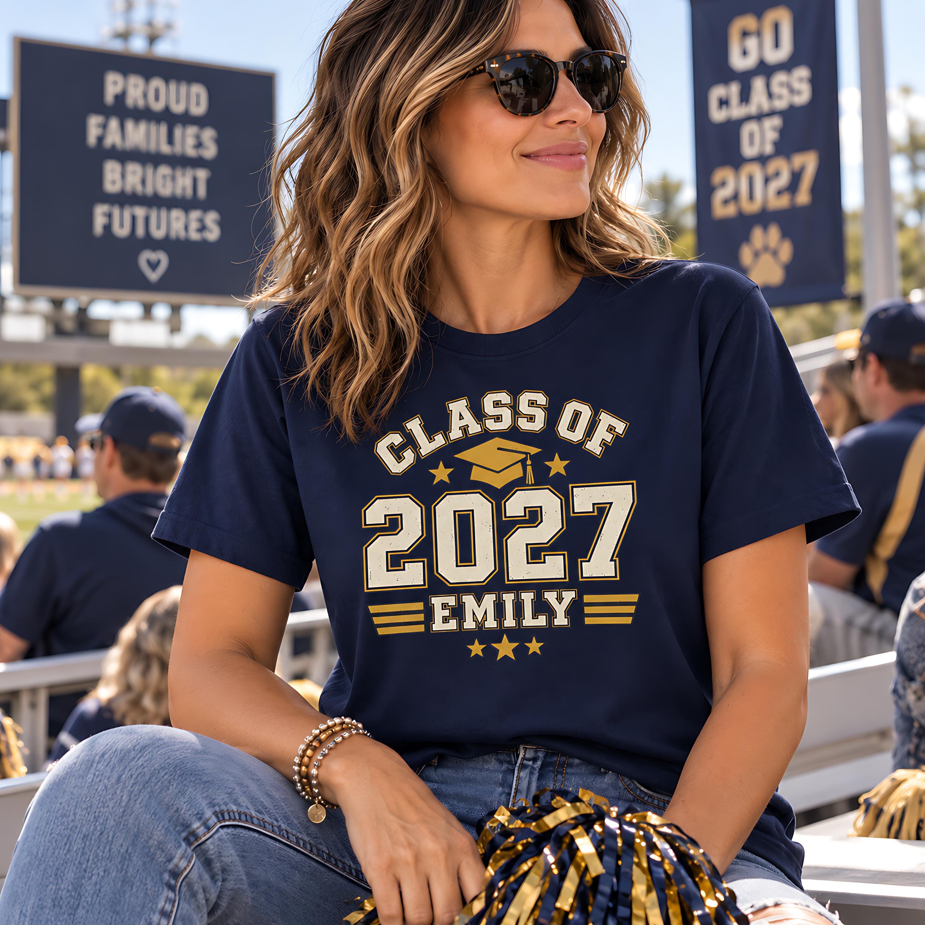 A woman wearing a navy blue t-shirt with the text "CLASS OF 2027 EMILY" is sitting in a stadium, smiling and holding a pom-pom.